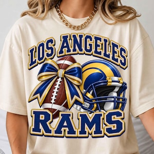 Może przedstawiać: Kremowy t-shirt z grafiką przedstawiającą napis LOS ANGELES RAMS w kolorze niebieskim i złotym. Wzór zawiera brązową piłkę do futbolu amerykańskiego ozdobioną dużą niebiesko-złotą kokardą oraz niebiesko-żółty kask futbolowy z maską ochronną.