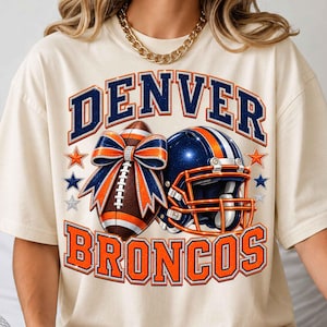Puede incluir: Una camiseta color crema con un estampado gráfico que incluye el texto DENVER BRONCOS en azul marino y naranja. El diseño presenta un balón de fútbol americano con un lazo grande azul y naranja, un casco azul y varias estrellas pequeñas.