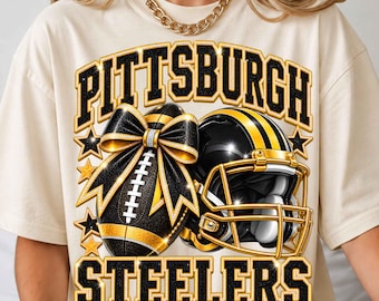 May include: A cream-colored t-shirt featuring a graphic of a black and gold football with a bow and a matching football helmet. The text PITTSBURGH STEELERS is printed in large black and gold block letters above and below the sports equipment.