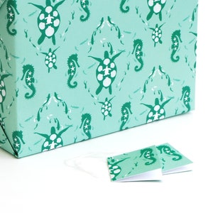 May include: A teal blue gift box with a repeating pattern of sea turtles and seahorses. The box is wrapped in paper with a white background. Two smaller teal blue gift tags with the same pattern are on a white surface.