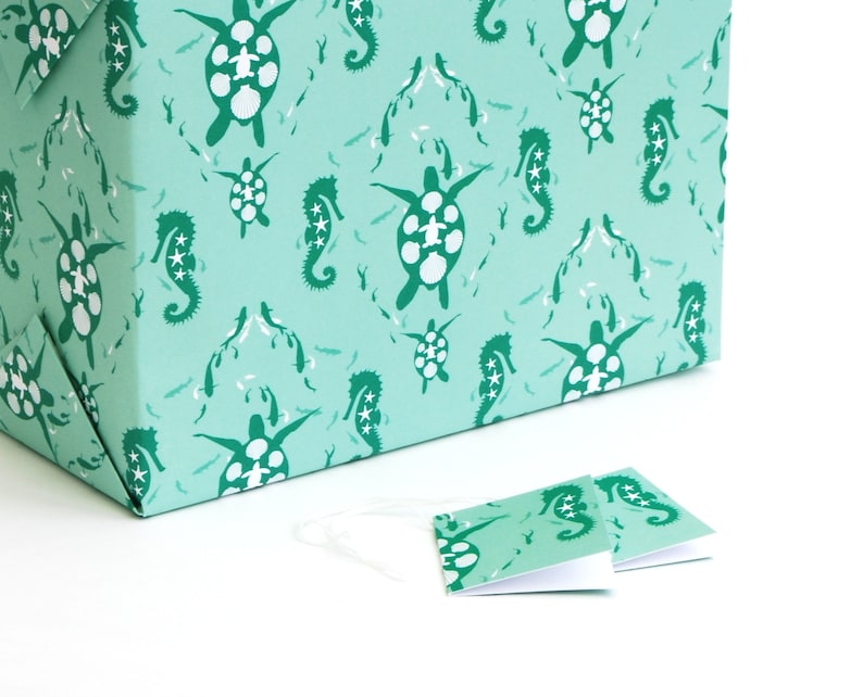 May include: A teal blue gift box with a repeating pattern of sea turtles and seahorses. The box is wrapped in paper with a white background. Two smaller teal blue gift tags with the same pattern are on a white surface.