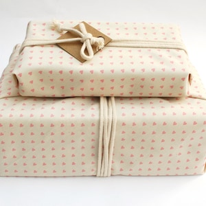 May include: Two gifts wrapped in white fabric with a pink heart pattern. The gifts are tied with twine and a brown paper tag.
