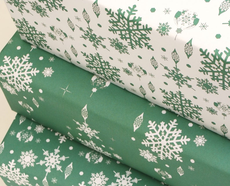 K&ouml;nnte beinhalten: Nahaufnahme von drei Geschenkpapierrollen. Die obere Rolle ist wei&szlig; mit einem gr&uuml;nen Schneeflockenmuster. Die mittleren und unteren Rollen sind gr&uuml;n mit einem wei&szlig;en Schneeflockenmuster. Das Papier ist aufgerollt und zeigt das Muster an den R&auml;ndern.