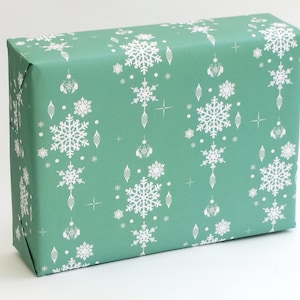 K&ouml;nnte beinhalten: Ein rechteckiger Geschenkkarton, der in t&uuml;rkisfarbenes Papier eingewickelt ist. Das Papier ist mit wei&szlig;en Schneeflocken, Sternen und dekorativen Ornamenten gemustert. Das Papier ist sauber um die Schachtel gefaltet.