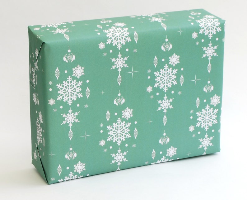 K&ouml;nnte beinhalten: Ein rechteckiger Geschenkkarton, der in t&uuml;rkisfarbenes Papier eingewickelt ist. Das Papier ist mit wei&szlig;en Schneeflocken, Sternen und dekorativen Ornamenten gemustert. Das Papier ist sauber um die Schachtel gefaltet.