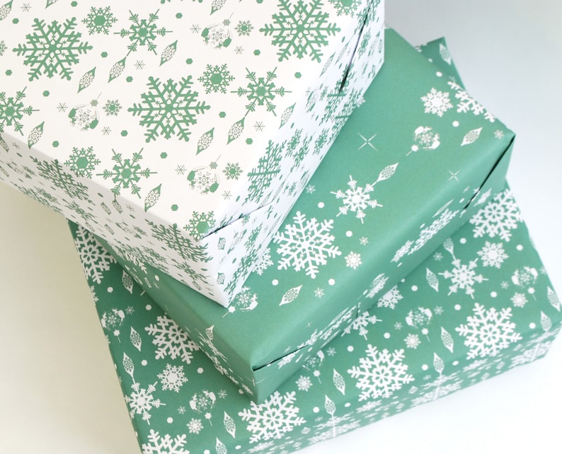 K&ouml;nnte beinhalten: Drei gestapelte Geschenkboxen, verpackt in festlichem Papier. Das Geschenkpapier zeigt ein sich wiederholendes Muster aus wei&szlig;en Schneeflocken und dekorativen Elementen auf gr&uuml;nem und wei&szlig;em Hintergrund. Die Boxen sind ordentlich gefaltet und bereit zum Verschenken.