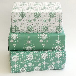 K&ouml;nnte beinhalten: Drei gestapelte Geschenkboxen, verpackt in festlichem Papier. Die obere Box zeigt wei&szlig;e Schneeflocken auf wei&szlig;em Hintergrund. Die mittlere Box ist in gr&uuml;nes Papier mit wei&szlig;en Schneeflocken- und Ornamentmustern gewickelt. Die untere Box ist in gr&uuml;nes Papier mit wei&szlig;en Schneeflockenmustern gewickelt.