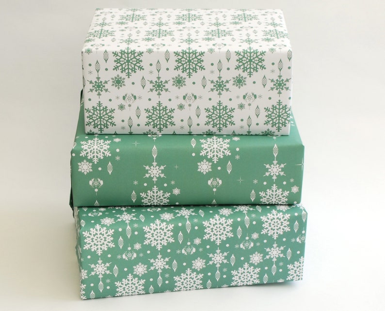 K&ouml;nnte beinhalten: Drei gestapelte Geschenkboxen, verpackt in festlichem Papier. Die obere Box zeigt wei&szlig;e Schneeflocken auf wei&szlig;em Hintergrund. Die mittlere Box ist in gr&uuml;nes Papier mit wei&szlig;en Schneeflocken- und Ornamentmustern gewickelt. Die untere Box ist in gr&uuml;nes Papier mit wei&szlig;en Schneeflockenmustern gewickelt.