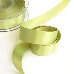 May include: A spool of light green satin ribbon. The ribbon is wound around a clear plastic spool and is lying on a white surface.