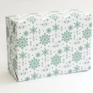 K&ouml;nnte beinhalten: Eine rechteckige Geschenkbox, verpackt in wei&szlig;em Papier mit einem sich wiederholenden Muster aus gr&uuml;nen Schneeflocken, Ornamenten und Punkten. Das Geschenkpapier ist sauber um die Box gefaltet, wodurch scharfe Kanten entstehen. Das Gesamtthema ist festlich und passend f&uuml;r die Weihnachtszeit.