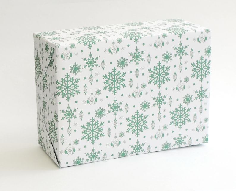 K&ouml;nnte beinhalten: Eine rechteckige Geschenkbox, verpackt in wei&szlig;em Papier mit einem sich wiederholenden Muster aus gr&uuml;nen Schneeflocken, Ornamenten und Punkten. Das Geschenkpapier ist sauber um die Box gefaltet, wodurch scharfe Kanten entstehen. Das Gesamtthema ist festlich und passend f&uuml;r die Weihnachtszeit.
