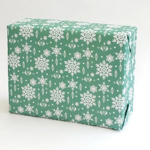 K&ouml;nnte beinhalten: Ein rechteckiger Geschenkkarton, verpackt in gr&uuml;nem Papier mit einem sich wiederholenden Muster aus wei&szlig;en Schneeflocken und dekorativen Ornamenten. Der Karton ist ordentlich gefaltet, ideal f&uuml;r die Weihnachtszeit.