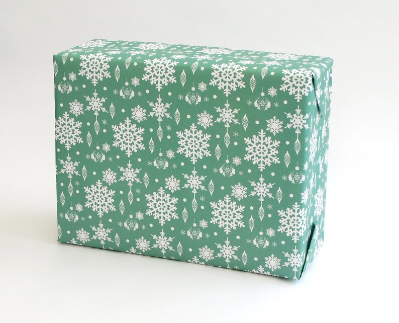 K&ouml;nnte beinhalten: Ein rechteckiger Geschenkkarton, verpackt in gr&uuml;nem Papier mit einem sich wiederholenden Muster aus wei&szlig;en Schneeflocken und dekorativen Ornamenten. Der Karton ist ordentlich gefaltet, ideal f&uuml;r die Weihnachtszeit.