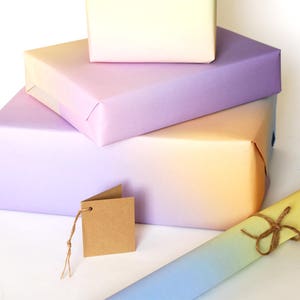 May include: A stack of three ombre gift wrap sheets in shades of purple, yellow and blue. The sheets are folded and stacked on top of each other. A brown gift tag is tied to the bottom sheet with twine.