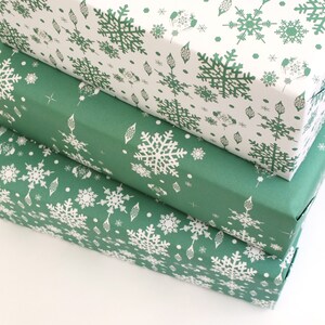 K&ouml;nnte beinhalten: Drei rechteckige Geschenkboxen, die &uuml;bereinander gestapelt sind. Die oberste Box ist wei&szlig; mit einem gr&uuml;nen Schneeflockenmuster. Die mittleren und unteren Boxen sind gr&uuml;n mit wei&szlig;en Schneeflockenmustern. Die Boxen sind in dekoratives Papier verpackt.