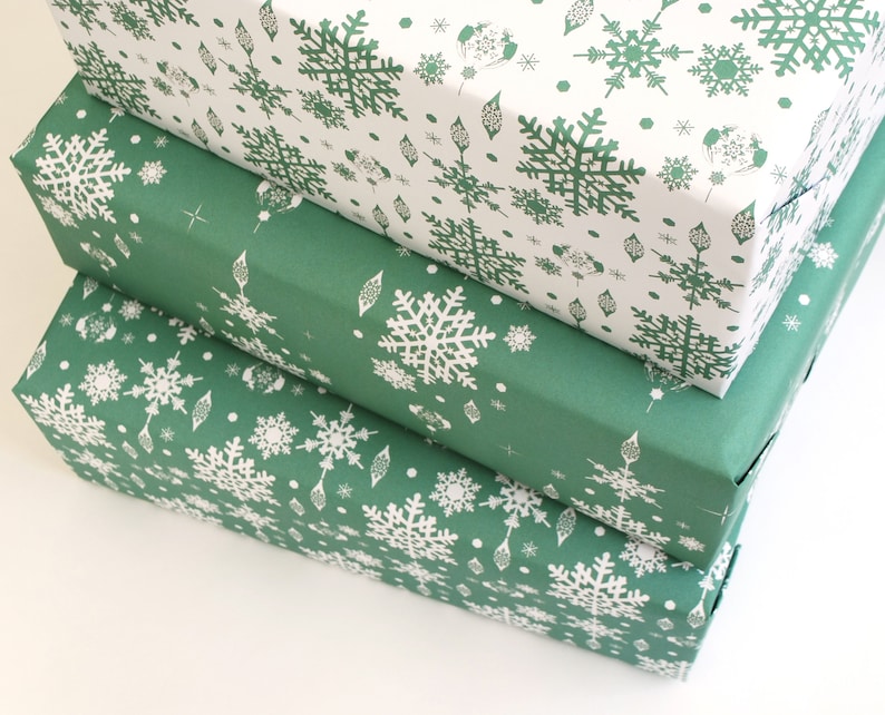 K&ouml;nnte beinhalten: Drei rechteckige Geschenkboxen, die &uuml;bereinander gestapelt sind. Die oberste Box ist wei&szlig; mit einem gr&uuml;nen Schneeflockenmuster. Die mittleren und unteren Boxen sind gr&uuml;n mit wei&szlig;en Schneeflockenmustern. Die Boxen sind in dekoratives Papier verpackt.