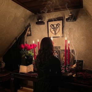 May include: A dark room featuring a wooden table with a black vase of red roses, an open book, and four tall red candles in black holders. A framed goat head illustration hangs on the wall above, with smoke rising from a metal incense burner nearby.