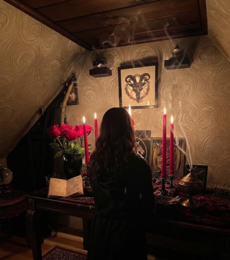 May include: A dark room featuring a wooden table with a black vase of red roses, an open book, and four tall red candles in black holders. A framed goat head illustration hangs on the wall above, with smoke rising from a metal incense burner nearby.