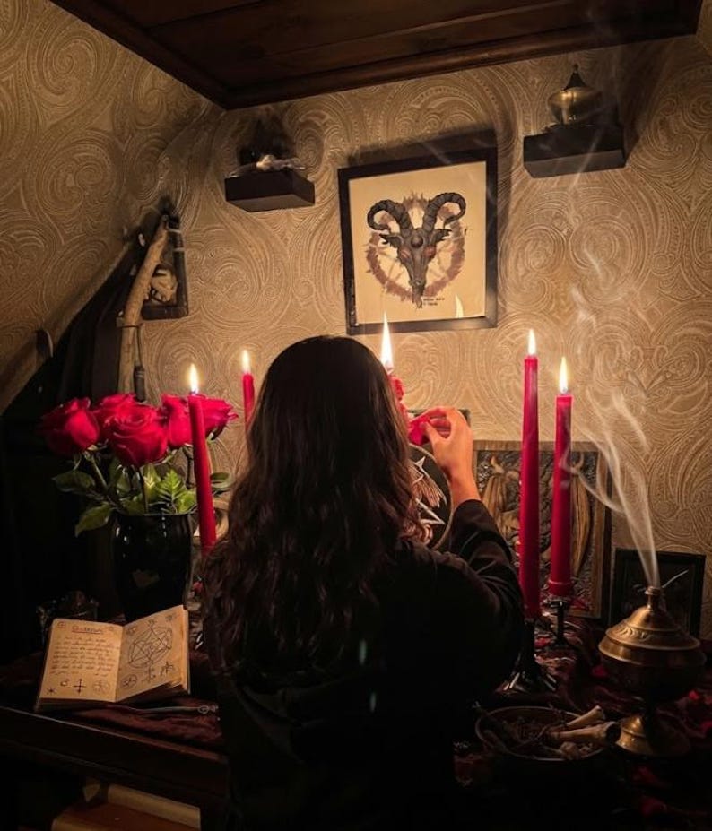 May include: A ritual altar setup featuring a framed goat skull illustration, four tall red taper candles, a bouquet of red roses in a black vase, an open book with handwritten text and geometric symbols, and a brass incense burner with rising smoke.