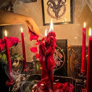 May include: A red sculptural candle shaped like two figures embracing, set in a dark metal tray. The candle is lit, surrounded by four tall red taper candles and scattered red rose petals. Smoke swirls around the scene, which includes framed artwork.
