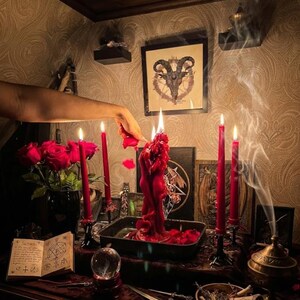 May include: A red sculptural candle shaped like two figures embracing, surrounded by four tall red taper candles in black holders. A bouquet of red roses, an open book with occult symbols, a crystal ball, a metal dagger, and incense smoke are present.