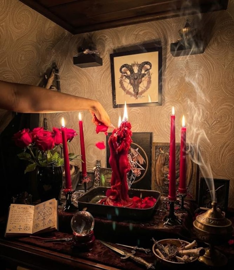 May include: A red sculptural candle shaped like two figures embracing, surrounded by four tall red taper candles in black holders. A bouquet of red roses, an open book with occult symbols, a crystal ball, a metal dagger, and incense smoke are present.