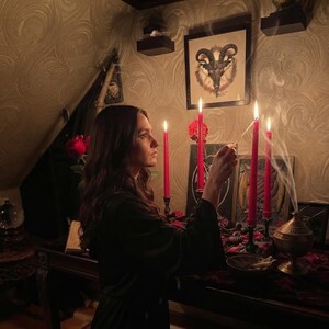 May include: A person in a black robe lighting a red candle with a match. The scene includes four tall red candles in black holders, a brass incense burner emitting smoke, a framed goat skull illustration, and red roses on a dark wooden table.