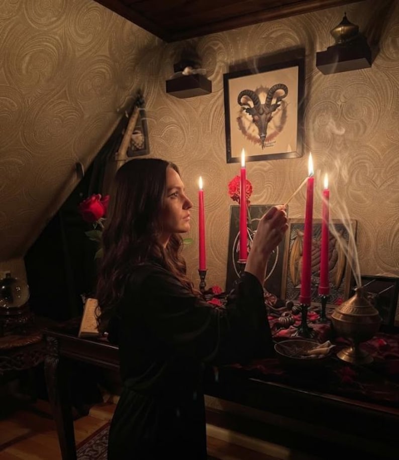 May include: A person in a black robe lighting a red candle with a match. The scene includes four tall red candles in black holders, a brass incense burner emitting smoke, a framed goat skull illustration, and red roses on a dark wooden table.