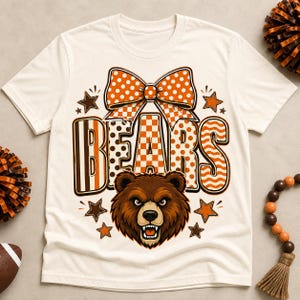 Può includere: Una t-shirt color crema con la grafica di una testa di orso bruno sotto la parola BEARS in grandi lettere decorate. Il design include un fiocco a pois arancione e bianco sopra il testo, circondato da piccole stelle arancioni e marroni.