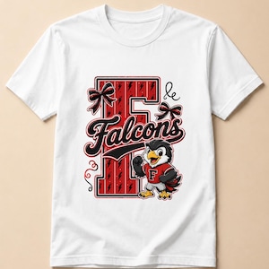 Op de afbeelding: Een wit t-shirt met korte mouwen voorzien van een grote rode letter F met zwarte bliksemschichtpatronen. Het ontwerp bevat het woord Falcons in zwarte schrijfletters, twee zwart-rode strikken en een valkenmascotte in een rood shirt met een F.