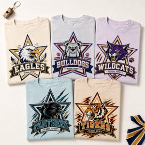 以下が含まれることがあります： クリーム、ライトグレー、ライトパープル、ライトブルー、タンのクルーネックTシャツ5枚。各シャツには、マスコットの頭部と「SCHOOL SPIRIT」の文字、そしてチーム名（EAGLES、BULLDOGS、WILDCATS、PANTHERS、TIGERS）が描かれた大きな星のグラフィックがプリントされています。