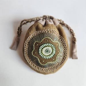 May include: A small, round, tan fabric drawstring pouch featuring a central embroidered floral motif in shades of green and cream. The pouch is cinched at the top with thick, braided tan yarn cords and displays intricate needlework detailing.