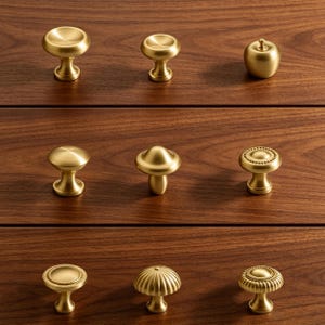 May include: A collection of nine brass cabinet knobs arranged in three rows. The knobs feature various shapes including smooth circular domes, a small apple, a mushroom-like form, and textured designs with beaded or fluted edges in a gold finish.