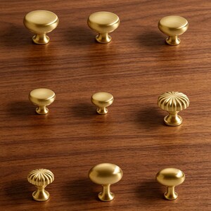 May include: A collection of nine brass cabinet knobs arranged in a grid. The set includes various shapes such as smooth circular domes, oval forms, and fluted designs, all featuring a consistent brushed metallic finish and a central mounting base.