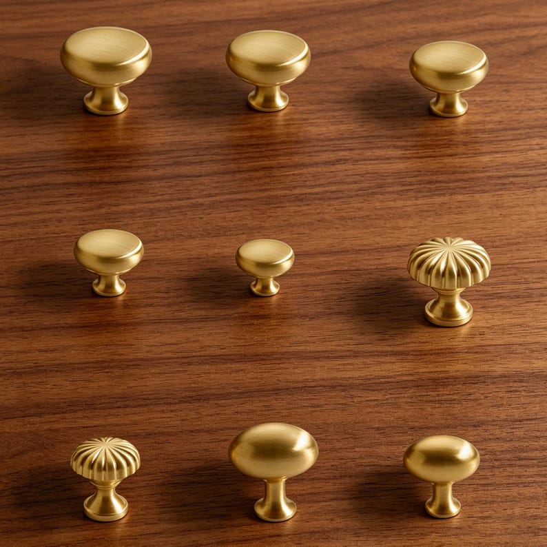 May include: A collection of nine brass cabinet knobs arranged in a grid. The set includes various shapes such as smooth circular domes, oval forms, and fluted designs, all featuring a consistent brushed metallic finish and a central mounting base.