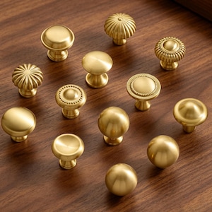 May include: A collection of twelve brass-toned cabinet knobs in various shapes, including smooth spherical, flat-topped, and ribbed designs. Each knob features a metallic finish and a circular base, showcasing a variety of textures and profiles.