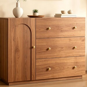 May include: A wooden cabinet with a light brown wood grain finish, featuring one vertical door with an arched panel design and three horizontal drawers. Each drawer and the door are equipped with a small, round, brushed gold metal knob.