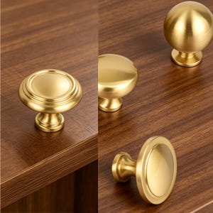 May include: A collection of three gold-colored metal cabinet knobs with a smooth, brushed finish. Each knob features a circular, rounded design with a central dome and a sturdy base, showcasing a consistent metallic luster across all pieces.