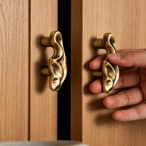 Può includere: Due maniglie per armadietto in ottone con una texture superficiale irregolare, organica e ondulata. Ogni maniglia è fissata a un'anta di armadietto in legno chiaro con due supporti cilindrici, caratterizzata da una finitura metallica.