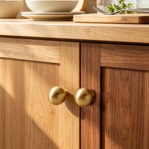 May include: Two spherical cabinet knobs with a smooth, brushed gold finish attached to light-toned wooden cabinet doors. The knobs are round and metallic, contrasting with the natural grain and warm brown texture of the wooden cabinetry surface.