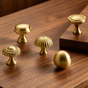 May include: Five brass cabinet knobs with various shapes including smooth domes, fluted designs, and textured edges. These metallic hardware pieces feature a brushed gold finish and are arranged in a group on a flat wooden surface.