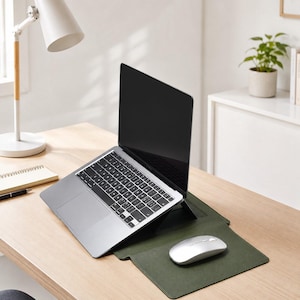 Könnte beinhalten: Ein silberner Laptop auf einem schwarzen, verstellbaren Ständer, platziert auf einer dunkelolivgrünen, rechteckigen Schreibtischunterlage. Eine silberne kabellose Computermaus liegt auf der rechten Seite der Unterlage. Der Laptop hat eine schwarze Tastatur.