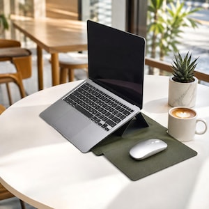 Könnte beinhalten: Ein silberner Laptop steht auf einem dunkelgrünen Klappständer und einer Schreibtischunterlage. Eine silberne kabellose Maus liegt auf der Unterlage neben dem Laptop. Eine weiße Keramiktasse mit Kaffee und eine kleine grüne Sukkulente in einem Marmortopf stehen daneben.