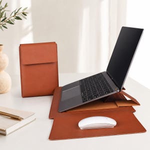 May include: A tan leather laptop stand that unfolds into a flat desk mat, featuring a dark grey laptop resting on the elevated portion and a white wireless mouse placed on the lower mat section, accompanied by a matching tan leather laptop sleeve.