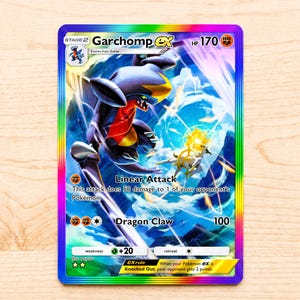 Peut inclure: Une carte à collectionner Garchomp ex avec une bordure holographique arc-en-ciel. La carte montre le personnage Garchomp dans une pose dynamique avec des couleurs bleu, noir et rouge, ainsi que le texte Linear Attack et Dragon Claw avec 170 HP.