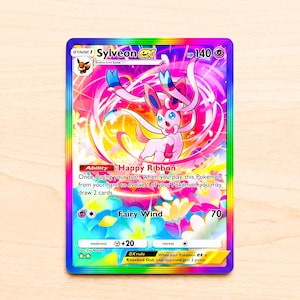 Könnte beinhalten: Eine holografische Sylveon EX Sammelkarte mit einem leuchtenden Regenbogenrand und einer zentralen Illustration des rosa Wesens, umgeben von wirbelnder Energie und bunten Blumen. Die Karte zeigt 140 KP, die Fähigkeit Happy Ribbon und den Angriff Fairy Wind.