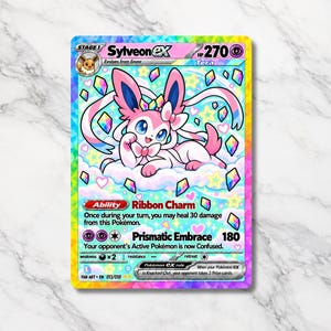 Puede incluir: Una carta coleccionable Sylveon ex que muestra una criatura rosa y blanca descansando sobre una nube. La carta indica 270 HP, la habilidad Ribbon Charm y el ataque Prismatic Embrace. El borde tiene un patrón de arcoíris con formas de cristal.