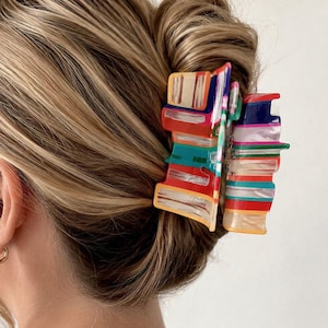 Puede incluir: Una pinza de pelo rectangular con un vibrante patrón de rayas multicolor en tonos rojo, azul, verde, naranja y blanco. El accesorio tiene un acabado brillante y un mecanismo de bisagra resistente diseñado para sujetar el cabello firmemente.