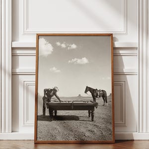 May include: A framed sepia-toned photograph depicting a person wearing a cowboy hat and suit leaning over a pool table in a desert landscape. A saddled horse stands nearby on the dry ground under a bright sky with scattered clouds in the distance.