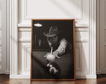 May include: A black and white framed wall art print depicting a person wearing a cowboy hat whilst playing billiards. The subject is leaning over a pool table, aiming a cue stick at a white cue ball positioned directly in front of the black eight ball.
